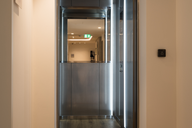 Modern stainless-steel elevator cab in a narrow hallway; doors closed, warm interior lighting, exit sign visible through the doorway ahead.