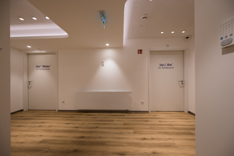 Interior corridor with two white doors labeled Suite 1 and Suite 2, warm wood floor and soft ceiling lights in a quiet resting area.