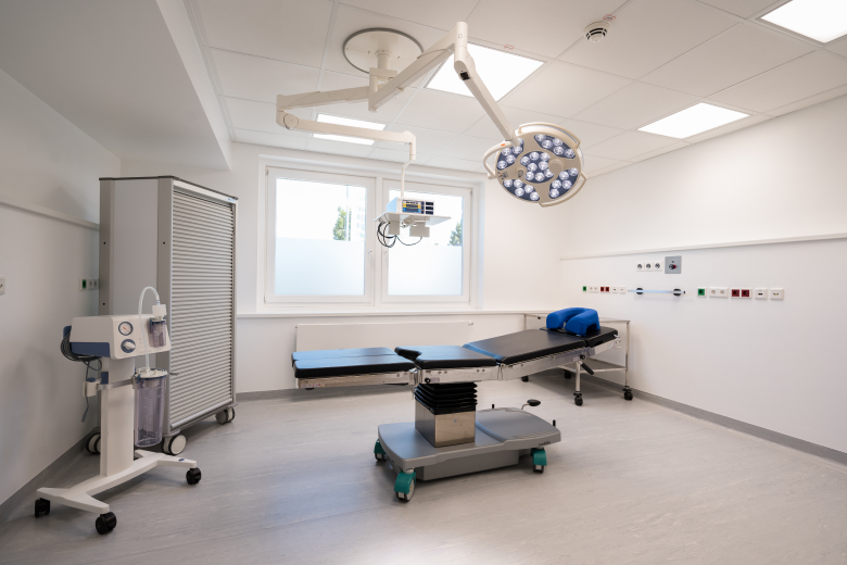 Modern operating room with an adjustable surgical table, overhead LED lights, and anesthesia equipment by the wall window.