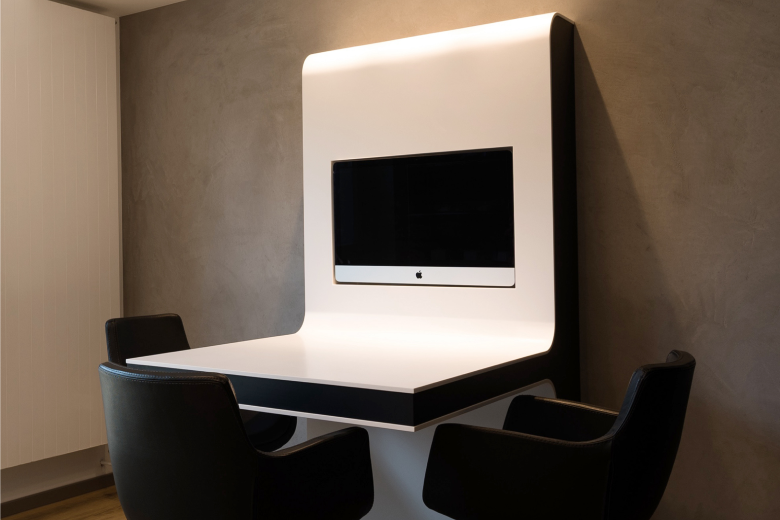 White curved desk with a mounted computer monitor in a small office nook, surrounded by four black chairs.
