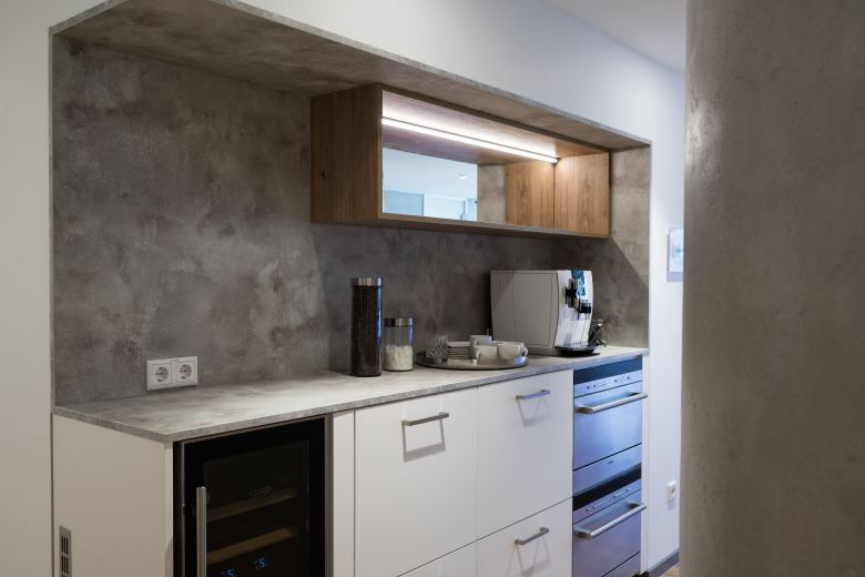 Modern kitchenette with white cabinets, concrete-textured wall, and built-in oven; coffee machine and canisters on the counter.