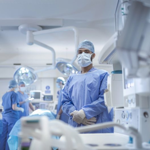 Surgeon in blue scrubs and mask stands beside the operating table in a bright, sterile operating room.